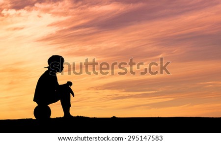 Silhouette of a boy sitting on football or soccer ball at the beach with sunset background. Childhood, serenity, sport, lifestyle concept