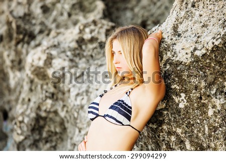 Young blonde girl in bikini leaning over a rock on sunny summer day. Beauty concept