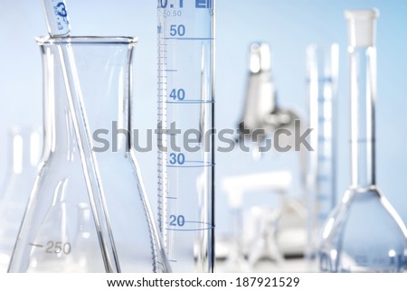 Close-up with Erlenmeyer flask, measuring jar and pipette with a microscope in the background.