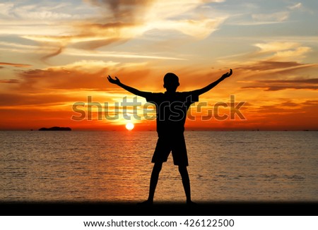 Silhouette of a boy with hands raised on a beach at the sunset concept for religion, worship, prayer and praise.