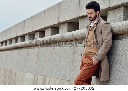 Handsome young man stands in a coat on the sand in windy weather