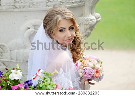 Beautiful sexy model girl in a white wedding dress. Beauty blonde bride without groom. Female portrait with bouquet. Woman with curly hair and lace veil in the park. Cute lady outdoors