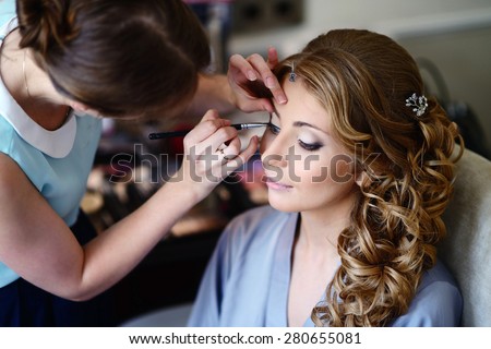 Wedding makeup artist making a make up for bride. Beautiful sexy model girl indoors. Beauty blonde woman with curly hair. Female portrait. Bridal morning of a cute lady. Close-up hands near face