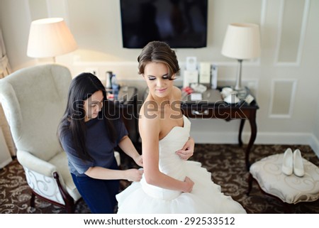 Bridesmaid is lacing white wedding dress for beautiful bride. Beauty model girl in bridal gown for marriage. Female portrait. Woman with curly hair. Cute lady indoors