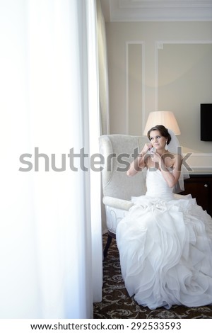 Beautiful bride in white wedding dress puts on earring. Beauty model girl is wearing jewelry. Female portrait in bridal gown for marriage. Woman with curly hair and lace veil. Cute lady indoors