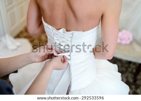 Bridesmaid is lacing white wedding dress for beautiful bride. Beauty model girl in bridal gown for marriage. Female portrait. Woman with curly hair. Cute lady indoors
