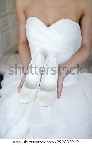 Beautiful bride in white wedding dress is holding shoes in her hands. Beauty model girl in bridal gown for marriage. Female portrait with boots. Close-up woman\'s arms. Cute lady indoors