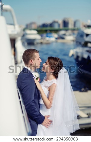 Wedding couple is hugging on a yacht. Beauty bride with groom. Beautiful model girl in white dress. Man in suit. Female and male portrait. Woman with lace veil. Cute lady and guy outdoors
