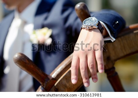 Groom is wearing a wrist watch indoors. Male portrait of handsome guy. Beautiful model boy in colorful wedding clothes. Man is posing