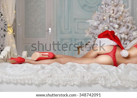 Beautiful sexy Santa Clause in elegant panties, hat and bra. Fashion portrait of model girl indoors with christmas tree. Cute woman in lace red lingerie. Female ass in underwear. Naked body