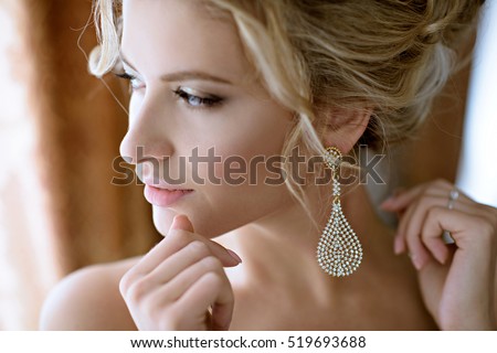 Beautiful bride in puts on earring. Beauty model girl is wearing jewelry for marriage. Wedding female portrait. Woman with curly hair and lace veil. Cute lady indoors