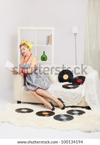 indoor portrait of beautiful young blonde size plus woman model sitting on sofa with vinyl records around and reading book