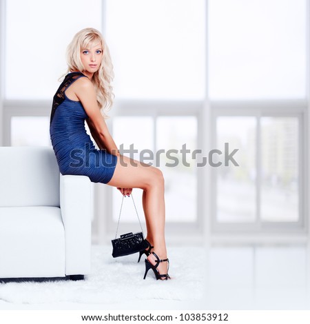 full-length portrait of beautiful young blond woman with handbag sitting on couch with white furs on floor