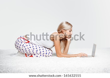 Young blonde woman in pyjamas sitting on white whole-floor carpet browsing laptop
