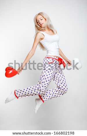 Young blonde woman in pyjamas jumping on gray background with red and white balloons in hands
