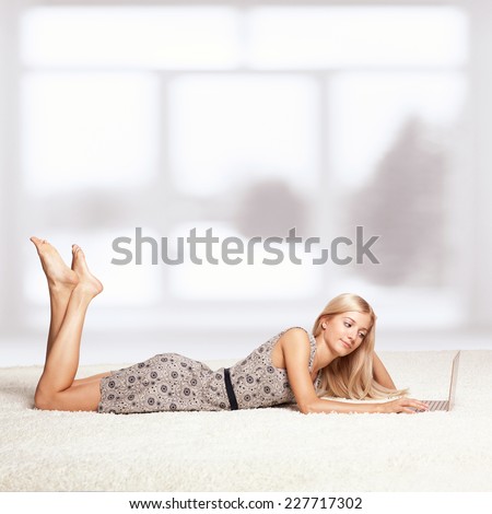 Blonde young woman on white whole-floor carpet browsing laptop  near window