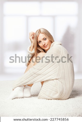 Blonde young woman dressed in long white cashmere sweater sitting on white whole-floor carpet and window  background
