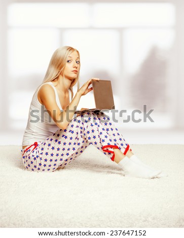 Young blonde woman in pyjamas on white whole-floor carpet browsing laptop  near window