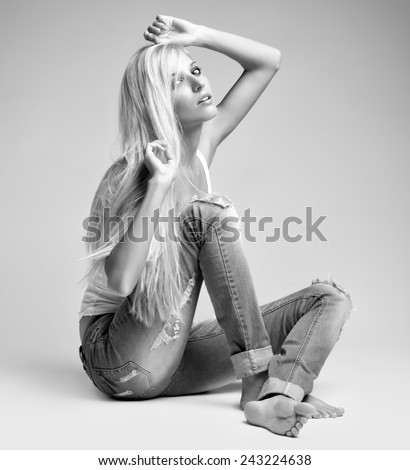 Monochrome portrait of blonde young woman in ragged jeans and vest sitting on floor on gray background