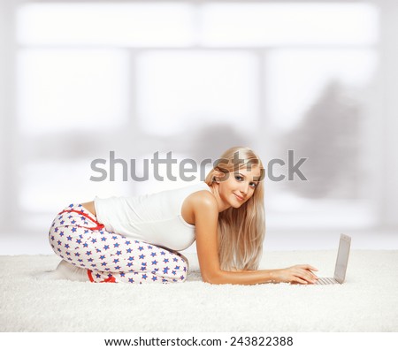 Young blonde woman in pyjamas sitting on white whole-floor carpet browsing laptop  near window