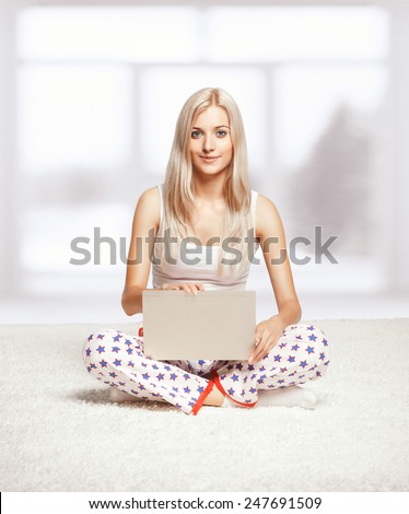 Young blonde woman in pyjamas sitting on white whole-floor carpet browsing laptop  near window
