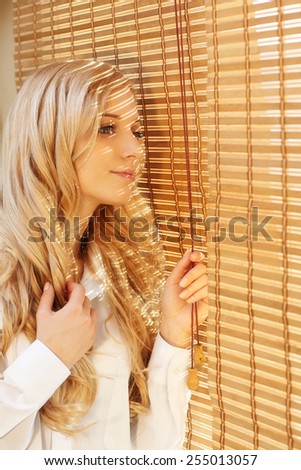 Young blonde beautiful happy woman looking out the window through the bamboo blinds with Sun hotspots on the face