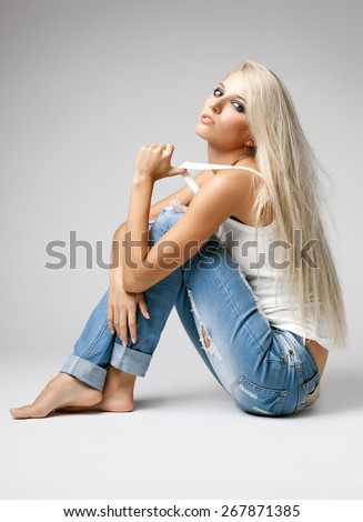 Blonde young woman in ragged jeans and vest sitting on floor on gray background