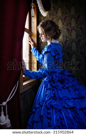 Young woman in blue vintage dress late 19th century standing near window in coupe of retro railway train