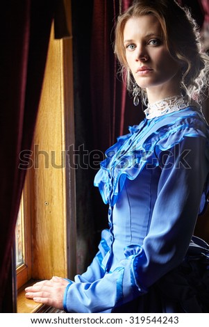 Young woman in blue vintage dress late 19th century standing near window in coupe of retro railway vehicle