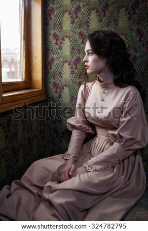 Young woman in beige vintage dress of early 20th century looking trough the window in coupe of retro railway train