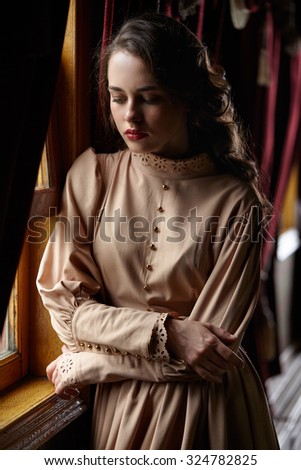 Young woman in beige vintage dress of early 20th century standing near window in corridor of retro railway train
