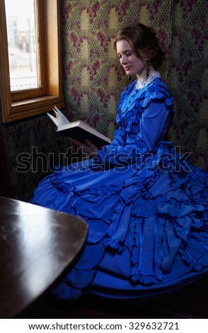 Young woman in blue vintage dress late 19th century reading the book in coupe of retro train