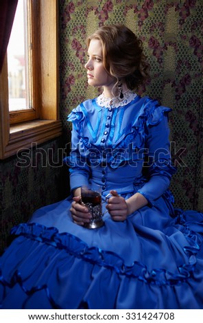 Young woman in blue vintage dress late 19th century sitting in coupe of retro railway train, looking out the window and drinking tea