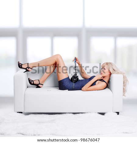 full-length portrait of beautiful young blond woman with hand bag lying on couch with white furs on floor