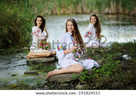 three girls in the Ukrainian national clothes with wreaths of flowers on the riverbank