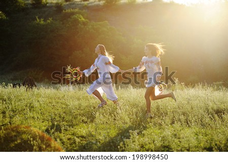 two girls in the national Ukrainian clothes with wreaths of flowers in their hands running over the grass
