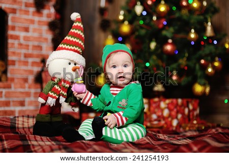 A child dressed as an elf sitting on a plaid background on the Christmas tree. Near the child in costume elf standing toy snowman