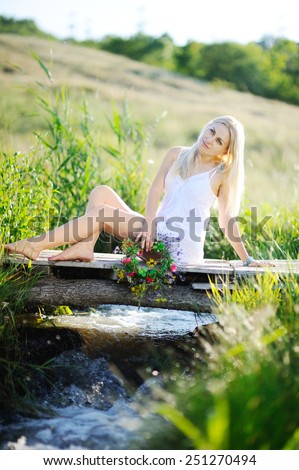 girl in a white sundress and with a wreath of flowers in hands sitting on the bridge. Stream flows under the bridge