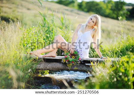 girl in a white sundress and with a wreath of flowers in hands sitting on the bridge. Stream flows under the bridge