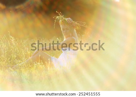girl in a white sundress and a wreath of flowers on her head sitting in the grass