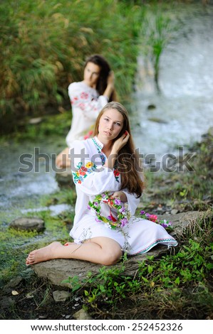 two girls in the Ukrainian national clothes with wreaths of flowers on the riverbank