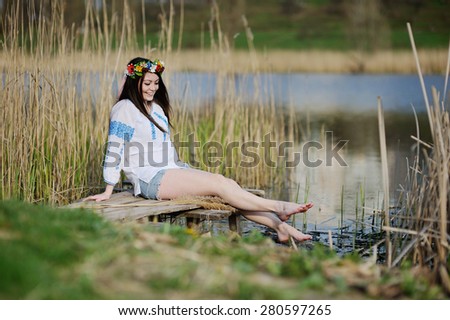 girl wets feet in the river. girl sitting on the bridge in the Ukrainian shirt and a wreath of flowers and ribbons on the head