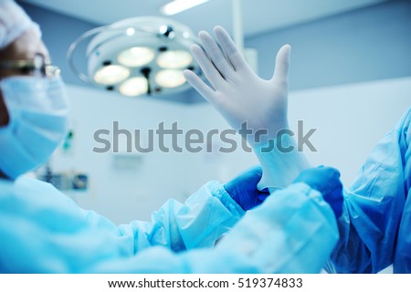 Assistant helps the surgeon put on latex gloves before the operation. A team of surgeons in the operating lamp background preparing for surgery