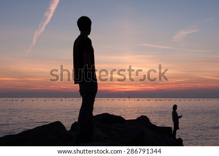 Silhouette of a man watching his friend fishing from rocks near the sea at sunset.