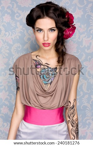 Young sensual attractive sexy european brunette girl with beautiful tattoo on her chest and arms posing for fashion photo shoot with red rose in her long hair in front of color floral background