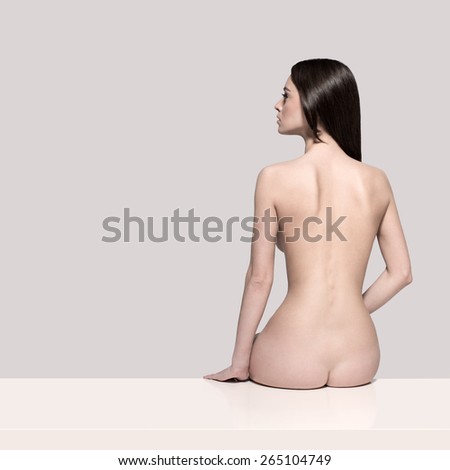 Young European fashion model woman with shiny healthy brunette hair, awesome gorgeous slim body and perfect skin is sitting on the table nude in studio for body care and wellness advertisement