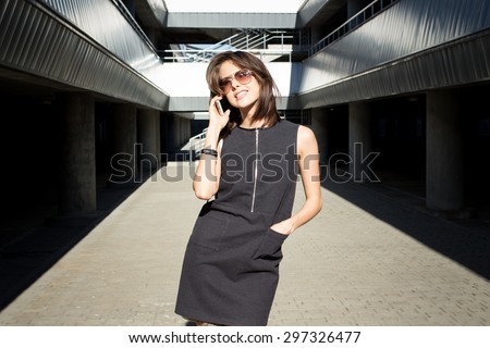 Attractive young brunette woman calling her model agency, holding mobile phone in her hand and standing outdoors in the city parking area on a bright sunny day