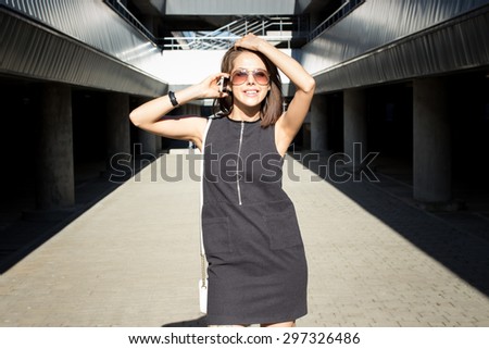 Attractive young brunette woman calling her model agency, holding mobile phone in her hand and standing outdoors in the city parking area on a bright sunny day