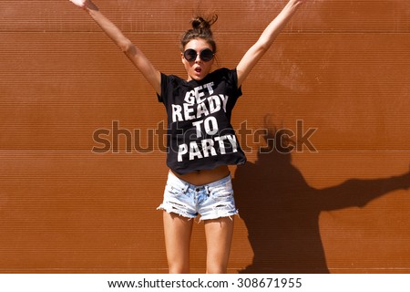 Young European teenage brunette curly model posing near brown ribbed wall, grimacing, jumping, wearing blue jeans mini shorts, sneakers, round sunglasses and black t-shirt, ready to party