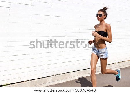 Young European teenage brunette sports model running outdoors along white wooden wall, jumping, jogging, wearing blue jeans mini shorts, sneakers, round sunglasses and black swimwear top